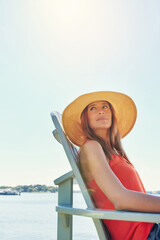 Fototapeta premium This is how I start everyday. a cheerful young woman wearing a hat while being seated on a chair next to a lake outside in the sun.