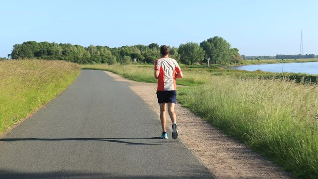 Steady moving shot of male trail runner seen from behind running on asphalt floodplains valley dyke in Dutch river IJssel landscape. Athlete endurance training run exercise outdoor sports workout.