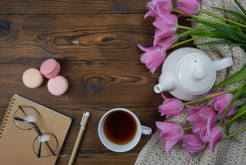 On the wooden table there is a white teapot and a cup of tea, a notebook and a bouquet of tulips