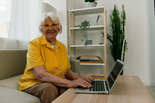 Happy Elderly Woman With A Laptop Typing With Headphones Sitting At Home On The Couch In A Yellow Shirt, Bright Modern Interior, Lifestyle Online Communication.