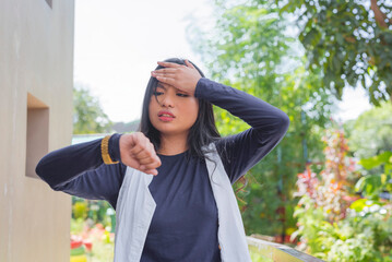Naklejka premium A close-up of a lovely lady in a cardigan and navy blue long-sleeved shirt, facing the camera with head up, palm on her forehead while checking her wrist watch.