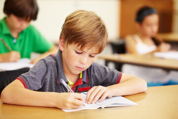 Boy kid, school classroom and writing test with focus, concentration and thinking for education goals. Male child, book and pen in class for assessment, studying or learning at desk for scholarship