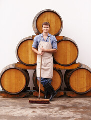 Portrait, broom and oak barrels with a man cleaning the wine cellar of a beverage distillery....