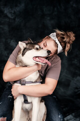 A young woman with blond hair hugs a husky dog on a dark background. The woman turned her face to the dog. Friendship between people and dogs. Caring for pets. Copy space