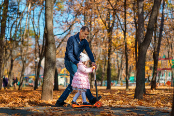 Fototapeta premium dad and child spend time together in autumn city park, happy people walk and relax, girl rides scooter, beautiful nature, bright sunny day and yellow leaves