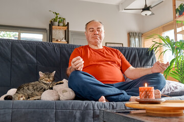 Overweight man practicing yoga and meditation at home sitting in lotus pose the couch, relaxed with closed eyes. Mindful meditation concept. Wellbeing.