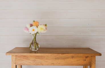 Colorful dahlia flowers in glass vase on oak side table against white panelled timber wall with copy space and vintage filter effect (selective focus)