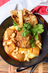 A serving of lamb shank Massaman curry, naan flatbread and potatoes in a dark round bowl
