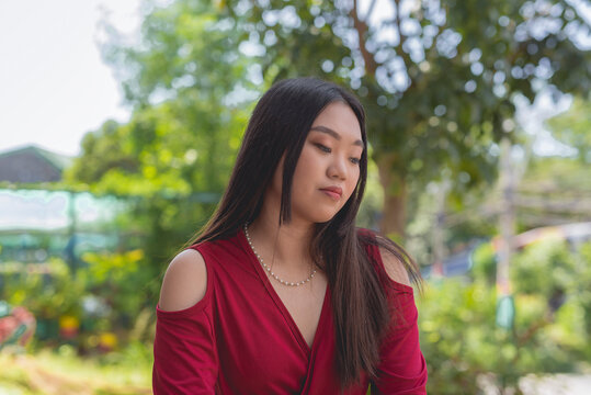 A close- up picture of a lovely white Asian woman in a vibrant red dress standing outside while head down and looking sad. Trees and plants in the background.