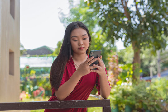 A white Asian Filipino girl in a long red dress leans against a rail with a serious expression, while focusing intently at her phone. Plants and trees are in the background.