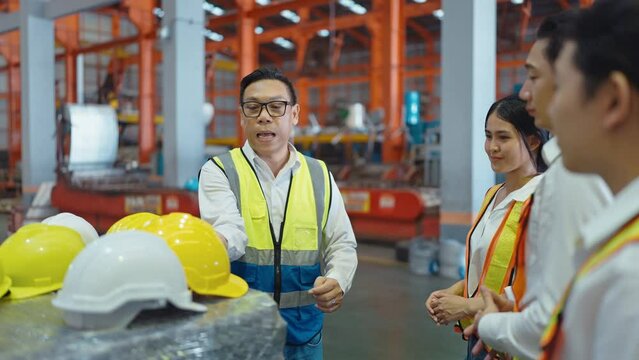 Asian male warehouse leader talking and recommend wearing helmets to team of warehouse workers and walking at industry manufacturing factory. Teamwork concept