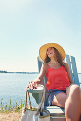 Fototapeta premium Nothing and nobody can spoil her mood. a cheerful young woman wearing a hat while being seated on a chair next to a lake outside in the sun.