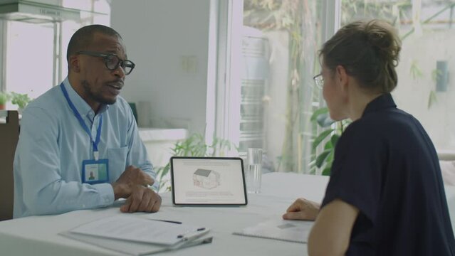 African American male engineer using digital table while presenting project of house powered with green energy to female client during meeting in office