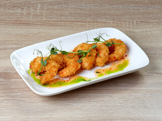Serving of five delicious tempura shrimp, decorated with micro greenery, in sweet and sour sauce, lying on a rectangular white plate, against the background of a wooden kitchen table. - 599514883