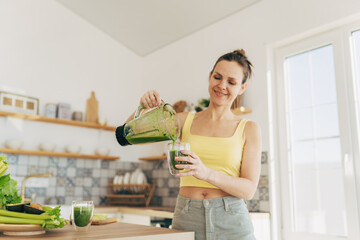 Happy woman on detox diet, pouring green cocktail from mixer into glass in kitchen