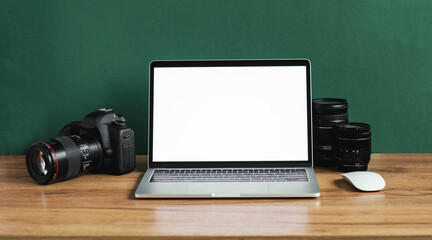 Modern photographer workplace with computer, DSLR camera and lenses. Laptop with the blank white screen on the wooden office table against dark green wall. Mockup for photo or videomaker advertisement