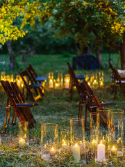 Candles in tall glass vases stand on the lawn. Wedding ceremony in the evening in the garden. Chairs in rows on the grass.