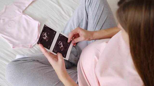 A young pregnant woman gently holds an ultrasound picture of her baby in her hands while sitting on the bed in the bedroom. Maternity concept.