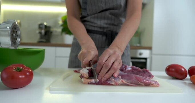Close-up view of woman cutting up fresh meat with a knife on a kitchen board.