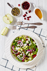 Green Salad with Apples, Goat Cheese, Cranberries, Red Onion and green pumpkin seeds in white bowl on white wood table with forks and ingredients, vertical view