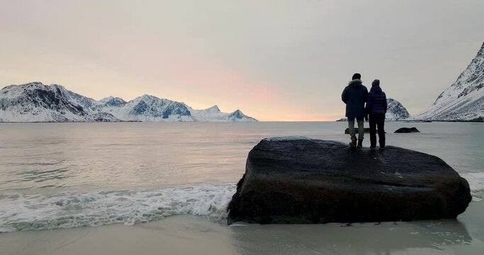 Travelers on rock look out into bay surrounded by snowy mountains, sunset aerial