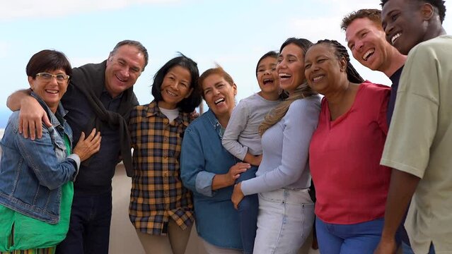 Group of multigenerational people hugging each other while smiling in front of camera - Multiracial friends having fun together outdoor
