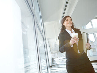 Business woman walking down a corridor holding a file and a coffee cup on her break. Happy mature...