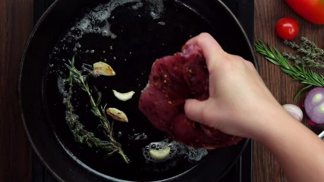 Hand puts an appetizing steak in a frying pan with rosemary, garlic and butter