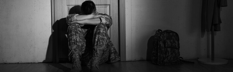 Black and white photo of depressed soldier sitting near backpack and door in hallway at home,...