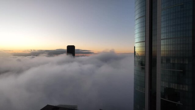 Aerial shot above a cloudy foggy Brisbane City, with only the tops of sky scrapers being visible. Sunrise. Flying nearby to a tall skyscraper. Camera facing Brisbane Skytower 222 Margaret St