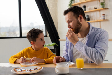 A boy in a yellow sweater is having breakfast in the kitchen with his father. Father and son eat sandwiches and look at each other.