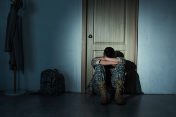 Lonely soldier in uniform sitting near backpack and door at home at night. - obrazy, fototapety, plakaty