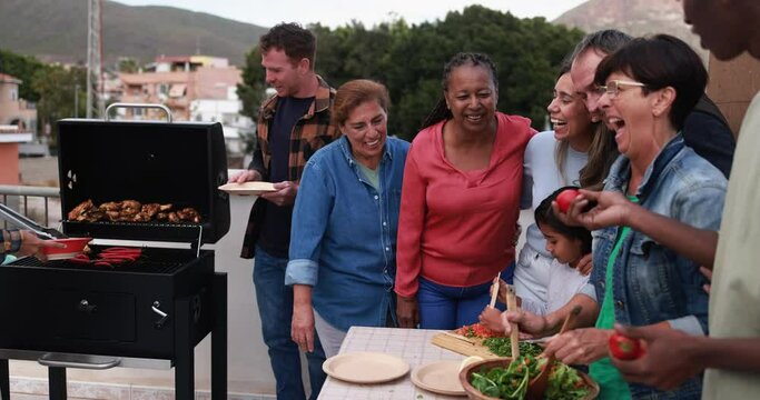 Multiracial friends having fun preparing food with child during weekend day - Multi generational people doing barbecue at home's rooftop