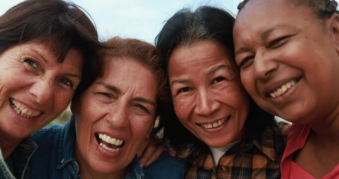 Joyful elderly women having fun hugging each other while smiling on camera - Multiracial female friends enjoy day together outdoor 