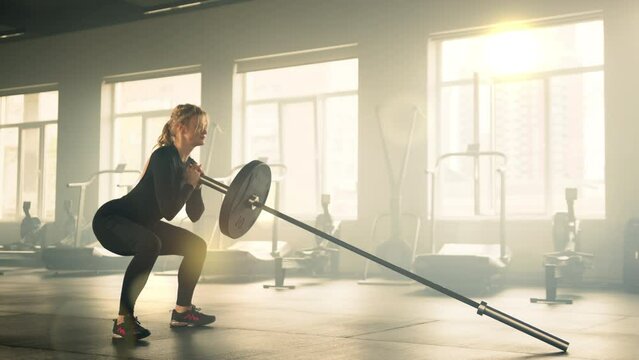 Profile shot of athletic young woman performing squats with barbell rod with one ball. Woman holds one end of barbell rod close to chest and squats. High quality 4k footage