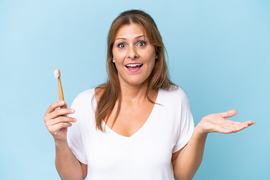 Middle-aged caucasian woman brushing teeth isolated on blue background with shocked facial expression