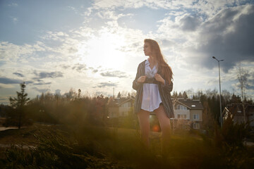 Naklejka na ściany i meble Beautiful girl with long hair in short skirt, white shirt and jacket in village or small town. Tall young slender woman and houses and sky with clouds on background on an autumn, spring or summer day