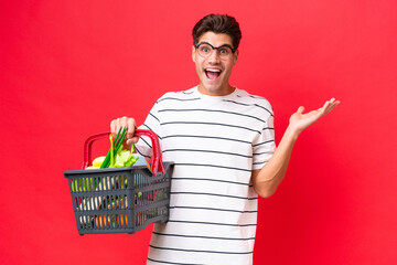 Young caucasian man man holding a shopping basket full of food isolated on red background with...