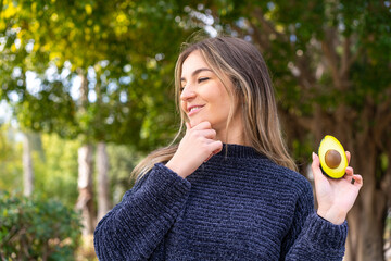 Young pretty Romanian woman holding an avocado at outdoors thinking an idea and looking side