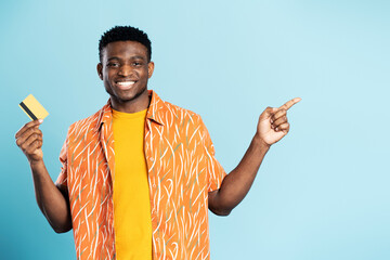 Handsome happy African American  man holding credit card looking at camera isolated on blue background, pointing on copy space. Shopping, electronic money, black Friday  concept