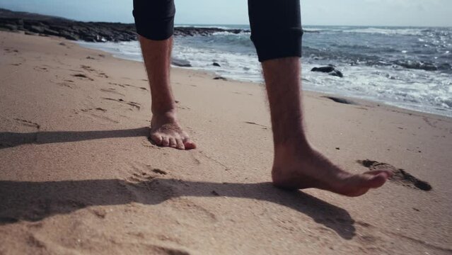 Close-up of bare guy feet walking along the sandy coast on a warm day. Legs of a walking man on a sandy beach against the background of the water, slow motion.