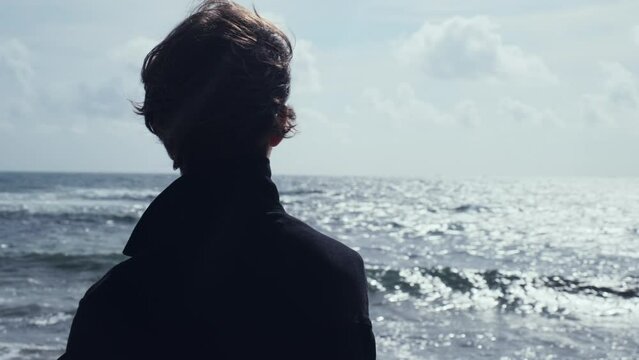 Young man stands on the empty sandy beach on the summer day and contemplating ocean water surface.  Human stands  on beach on a sunny day and looking to distance.  Back view of Man on the ocean shore