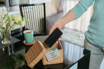 Close up of hand holding smartphone in wicker basket with inscription Device free zone. Woman putting phone into box with different gadgets at home. Digital detox and technology dependance concept