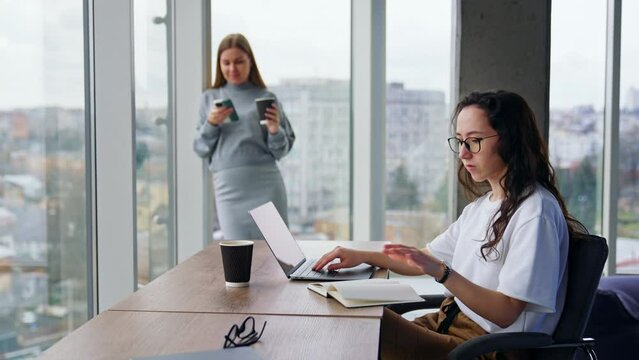 Businesswomen working in the modern office. Brunette woman typing on her laptop and blonde woman checks her phone .