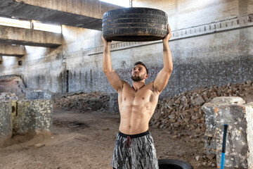 Strong ripped handsome man lifted a car tire above his head during fitness-cross fit workout exercise, factory complex background
