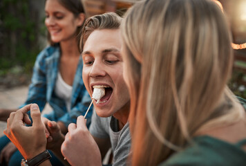 Open wide. a young cheerful man being fed a marshmallow by his girlfriend while being seated outside.