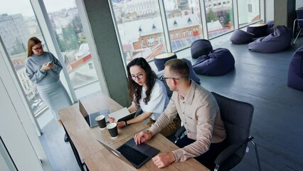People work in the modern light office. Man and woman sit at the desk looking on the laptop and communicating. Pregnant lady at the window uses her phone. High angle view. - Powered by Adobe
