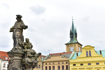Fototapeta na wymiar Ancient catholic sculptures on a pedestal and old buildings with green roofs and tower with the green top and clock. Statues on biblical subjects. Charles Bridge. Prague, Czech Republic, October 2022.