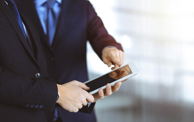 Business people use a tablet computer for discussion of their new project, standing in a sunny modern office. Unknown businessman or male entrepreneur with a colleague at workplace