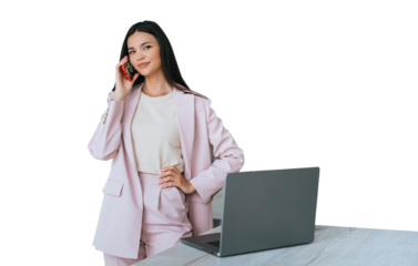 Satisfied brunette Asian young woman in pink suit talks by phone stands at desk with laptop looks at camera. Cheerful Korean businesswoman remote works home against transparent background.